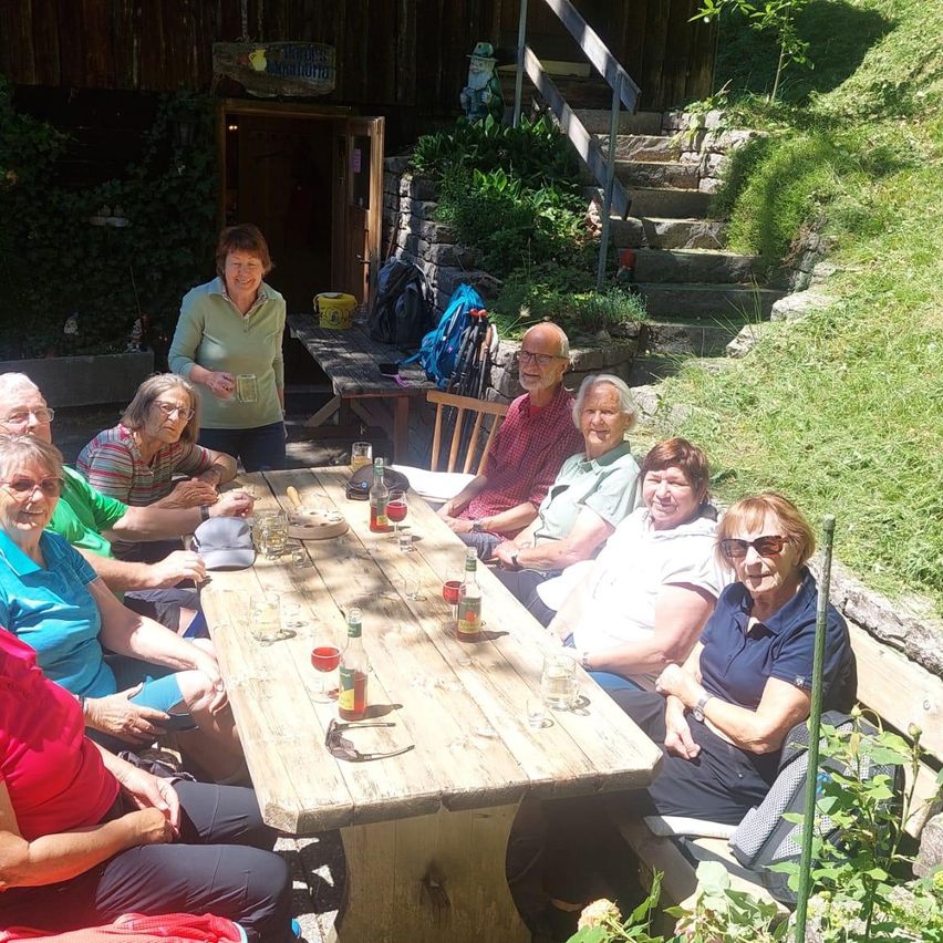 Eine Gruppe älterer Menschen sitzt draußen an einem Holztisch und genießt Getränke und Gespräche. Sie befinden sich in einem Garten mit Pflanzen und einer Treppe im Hintergrund.