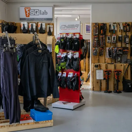 Ein Geschäft zeigt verschiedene Sportartikel, darunter Jacken, Handschuhe und Werkzeuge. Das Logo der Marke SQlab ist an einer Wand zu sehen.