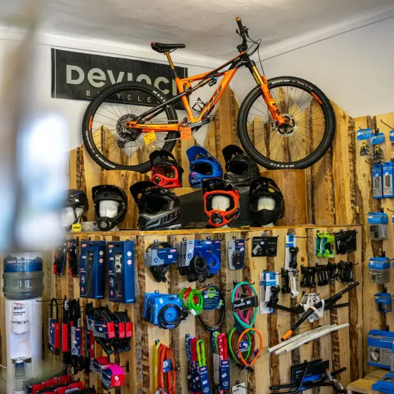 Ein Fahrradladen zeigt ein orangefarbenes Mountainbike, Helme und verschiedene Fahrradzubehörteile auf Holzregalen. Der Laden ist mit 'Devo' beschriftet.