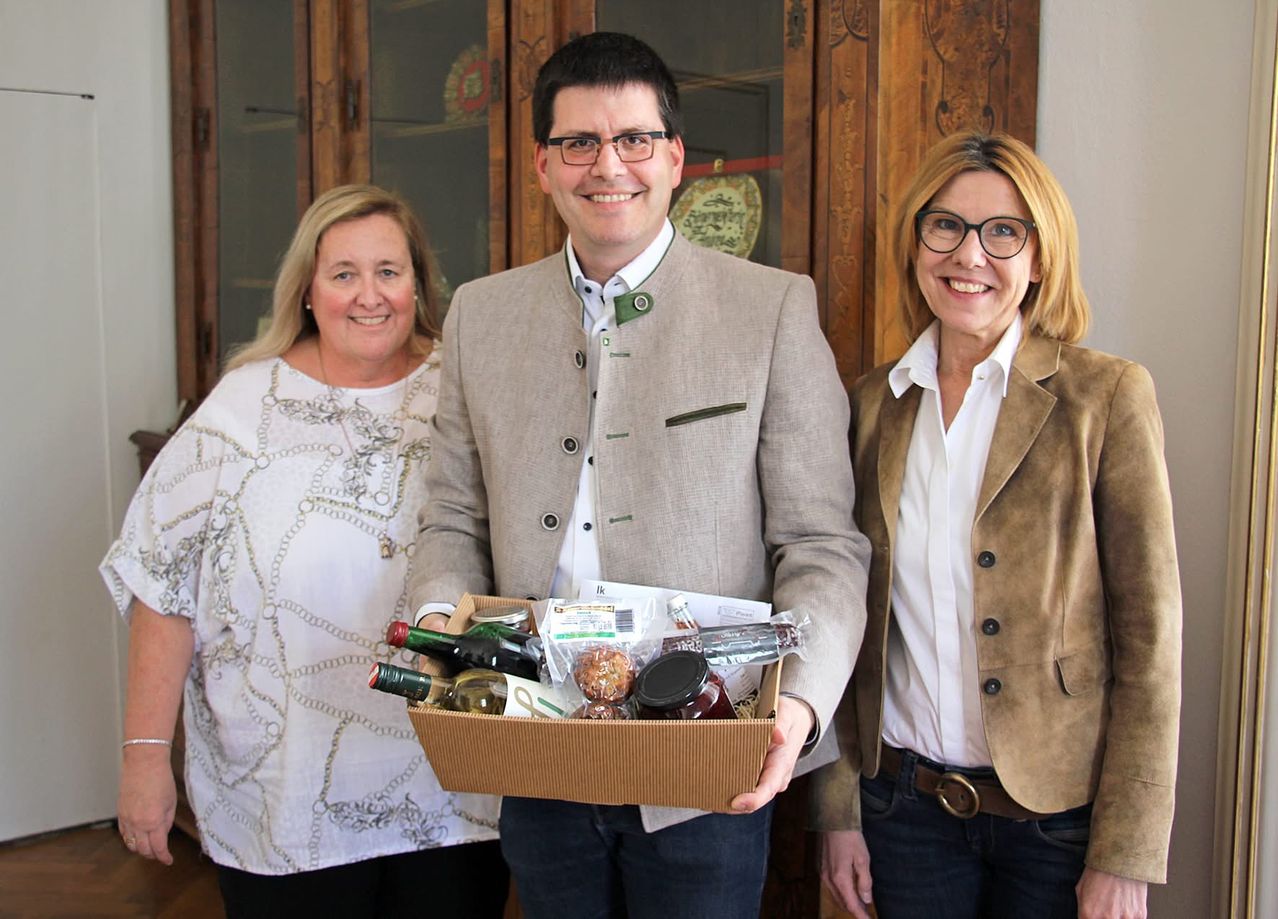 Drei Personen stehen nebeneinander und lächeln. Ein Mann hält eine Box mit Essen und Getränken. Hinter ihnen steht ein hölzerner Schrank mit Glastüren.