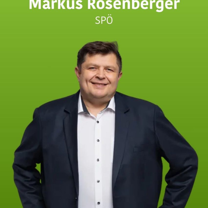 Ein lächelnder Mann, Markus Rosenberger, in einem weißen Hemd und dunkler Anzugjacke steht mit den Händen in den Hüften vor einem grünen Hintergrund.