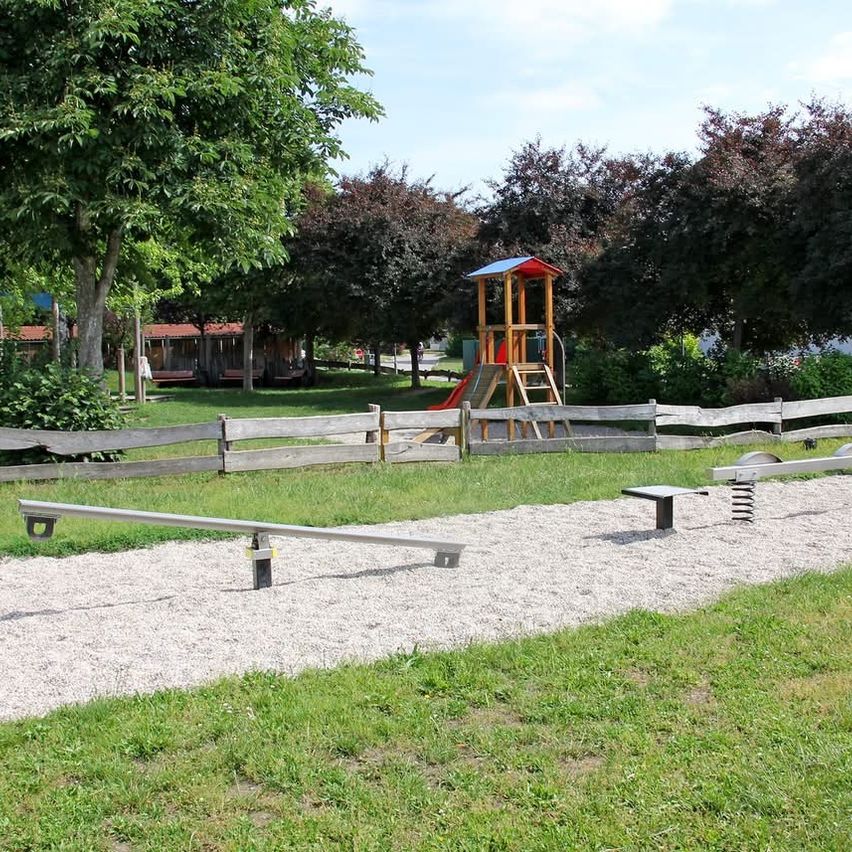 Ein Spielplatz mit einem roten Rutschen, einem hölzernen Klettergerüst und einem Zaun in einem Grasbereich mit Kies und Bäumen.