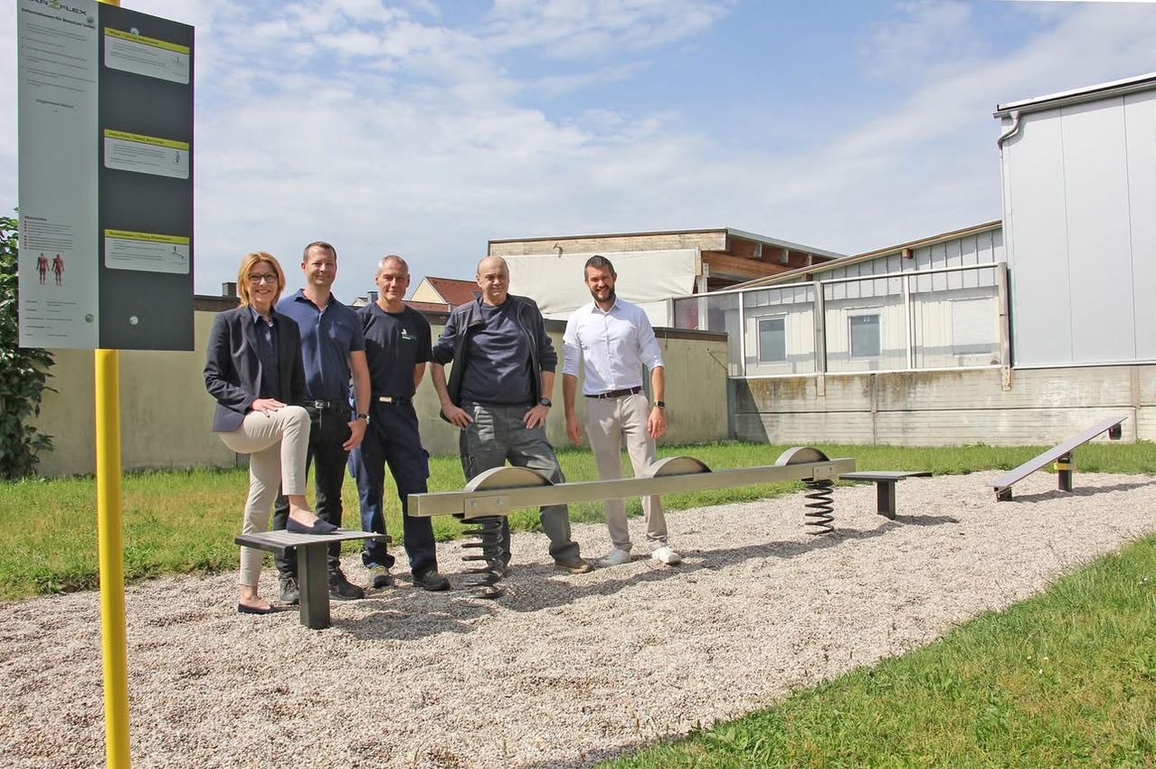 Fünf Personen stehen vor einem Wippgerät auf einem Kiesweg. Sie lächeln alle und scheinen für ein Foto zu posieren. Hinter ihnen befindet sich ein weißes Gebäude mit Fenstern.