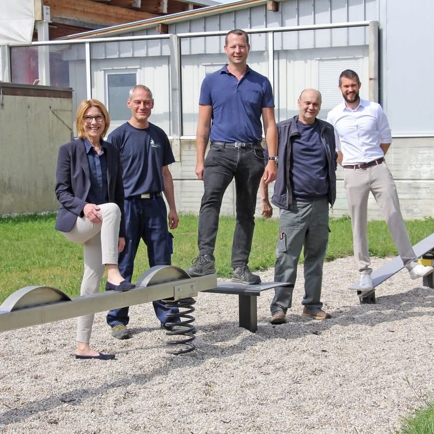 Fünf Personen stehen lächelnd auf einer Wippe im Freien. Ein Mann steht auf einer Feder, während die anderen auf der Wippe stehen. Hinter ihnen befindet sich ein Gebäude mit Glasfenstern.