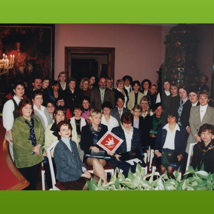 Eine Gruppe von Menschen, sowohl Männer als auch Frauen, steht und sitzt in einem Raum. Einige tragen Brillen, Halsketten und Schals. Eine Frau in der Mitte hält ein Buch. Eine Frau vorn hält ein Buch und einen Stift. Hinter ihnen sind Pflanzen. Links ist ein Gemälde eines Mannes. Rechts ist eine Lampe.