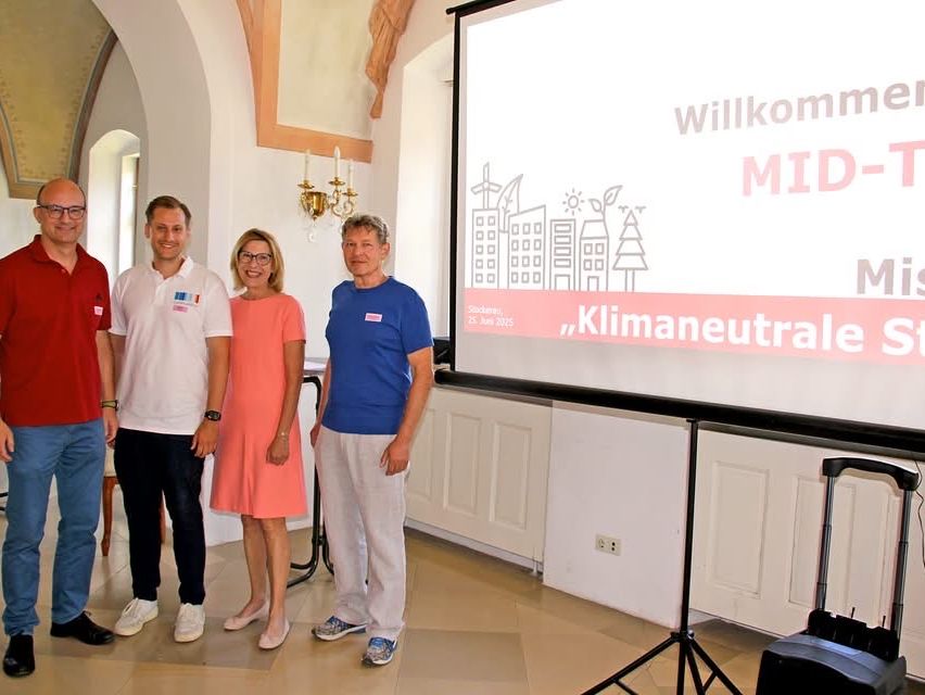 Vier Personen stehen in einem Raum mit einer Projektionsleinwand, auf der 'Willkommen MID-' und 'Klimaneutral' steht. Sie lächeln und scheinen für ein Foto zu posieren.