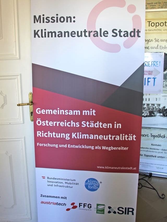 Ein Werbeposter für 'Mission: Klimaneutrale Stadt'. Es hat einen roten Hintergrund und Text über die Zusammenarbeit mit österreichischen Städten auf dem Weg zur Klimaneutralität. Es gibt Logos für das Bundesministerium für Innovation und Infrastruktur und AustriaTech.