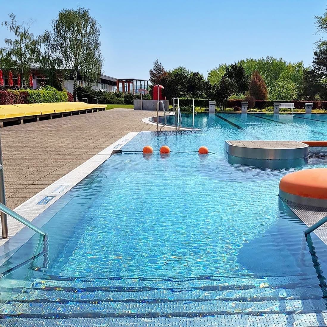 Ein Freibad mit einem blauen Poolbereich, einem Sprungbrett und orangefarbenen Schwimmringen. Der Pool hat Stufen und Handläufe auf beiden Seiten.