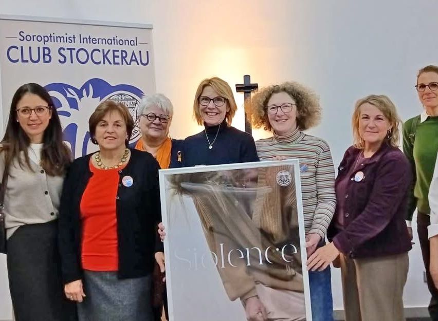 Eine Gruppe von Frauen posiert für ein Foto mit einem gerahmten Kunstwerk, das 'Silence' (Schweigen) besagt. Sie stehen hinter einem Banner für 'Optimist International Club Stockerau'.