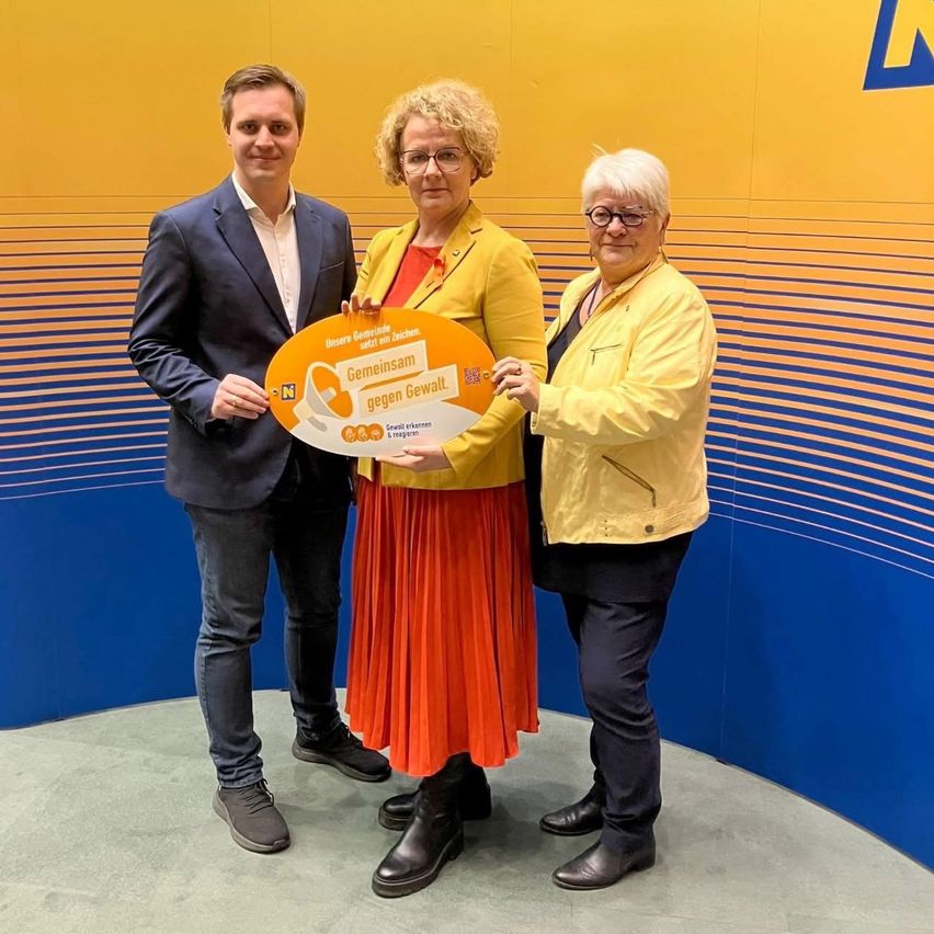 Drei Personen stehen zusammen und halten ein großes orangefarbenes Schild mit der Aufschrift 'Gemeinsam gegen Gewalt'. Zwei von ihnen sind Frauen und einer ist ein Mann. Sie stehen vor einer blau-gelben Wand.