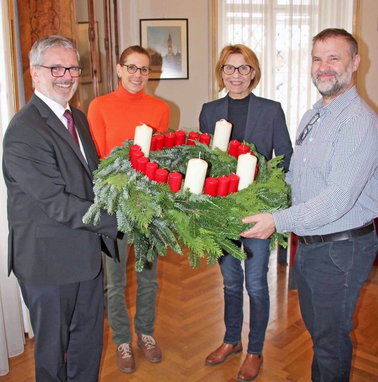 Vier Personen stehen zusammen und halten einen Kranz aus Grün und roten Kerzen, lächeln für ein Foto. Dahinter befindet sich ein Holzboden und ein Fenster mit Vorhängen.