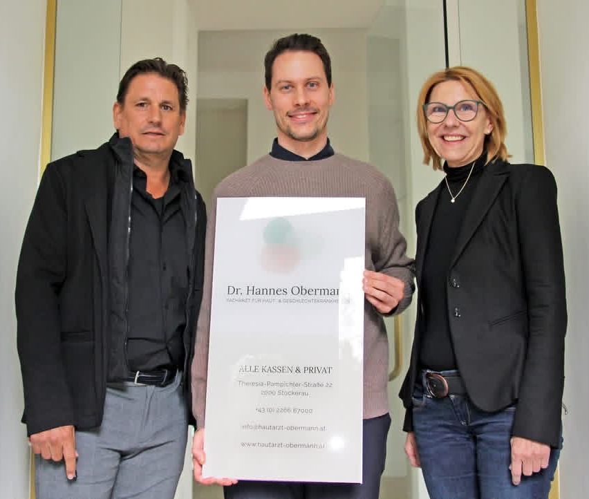 Drei Menschen stehen in einem Flur und halten ein Zertifikat für Dr. Hannes Obermann, einen Dermatologen. Der Mann in der Mitte hält das Zertifikat, während die anderen lächeln. Der Mann links trägt eine Jacke und Hose.