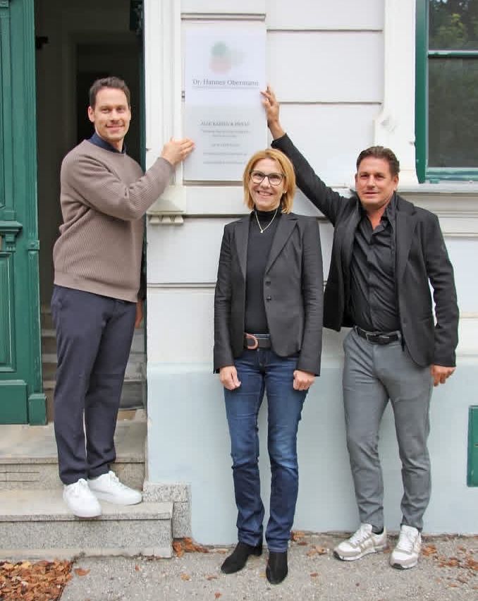 Drei Personen stehen vor einem Gebäude mit einem Schild, das 'Dr. Hanns Obermann' steht. Der Mann links trägt einen Pullover und Sneakers, die Frau in der Mitte hat eine Brille und eine Halskette, und der Mann rechts trägt einen Anzug und Lederschuhe.