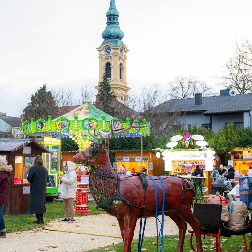 Ein Winterkarneval mit einem Karussell und einer roten Rentierstatue, mit Menschen und Ständen auf einer Grasfläche vor einem Turm.