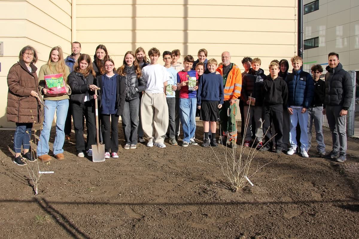 Eine Gruppe von Menschen, darunter Schüler und Erwachsene, posiert für ein Foto vor einem Gebäude. Einige halten Bücher, einer eine Schaufel. Sie alle lächeln.