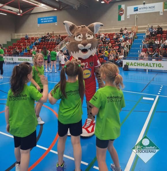 Fünf junge Mädchen in grünen Trikots stehen um ein Maskottchen in einem roten Trikot mit der Nummer 22 herum. Sie befinden sich auf einem Handballspielfeld.