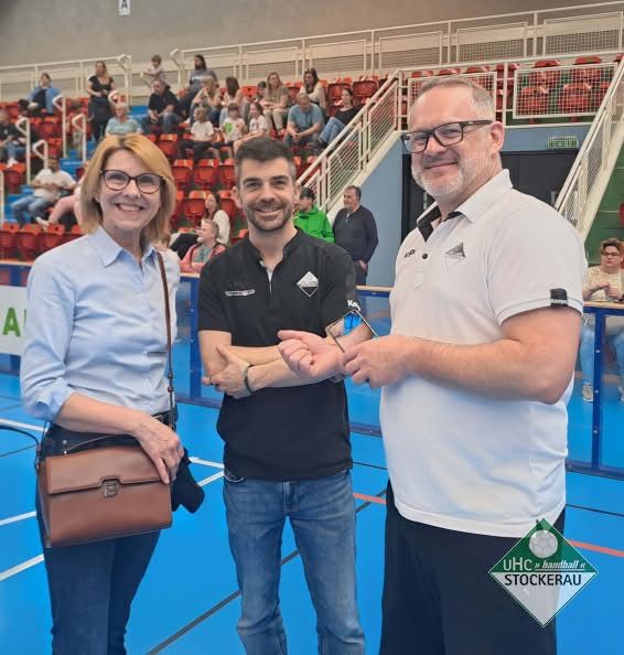 Drei Personen, zwei Männer und eine Frau, stehen in einer Sporteinrichtung. Die Frau lächelt und trägt eine braune Tasche. Der Mann auf der rechten Seite trägt eine Brille und ein Polohemd.