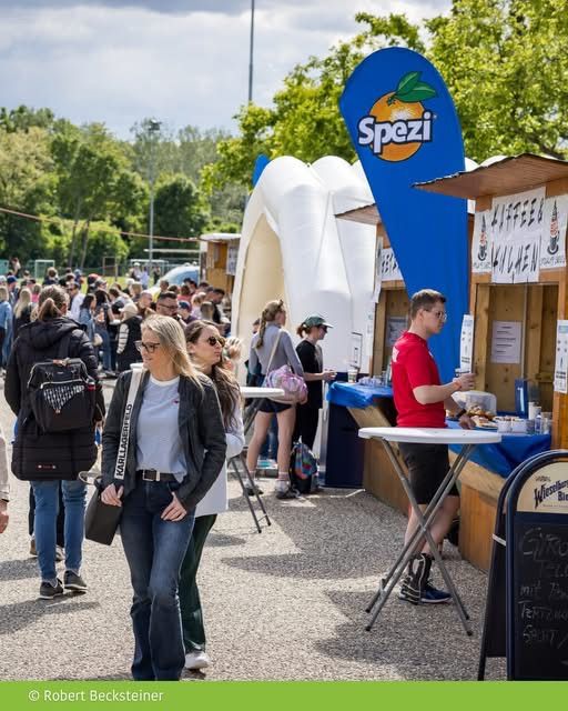 People walk past food stands in an outdoor area. A man in a red shirt serves food. A woman with a backpack looks at the food.