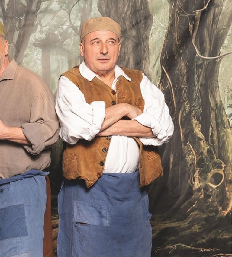 Two men in historical attire stand side by side in a forest setting. One man folds his arms while the other has his hands on his hips.