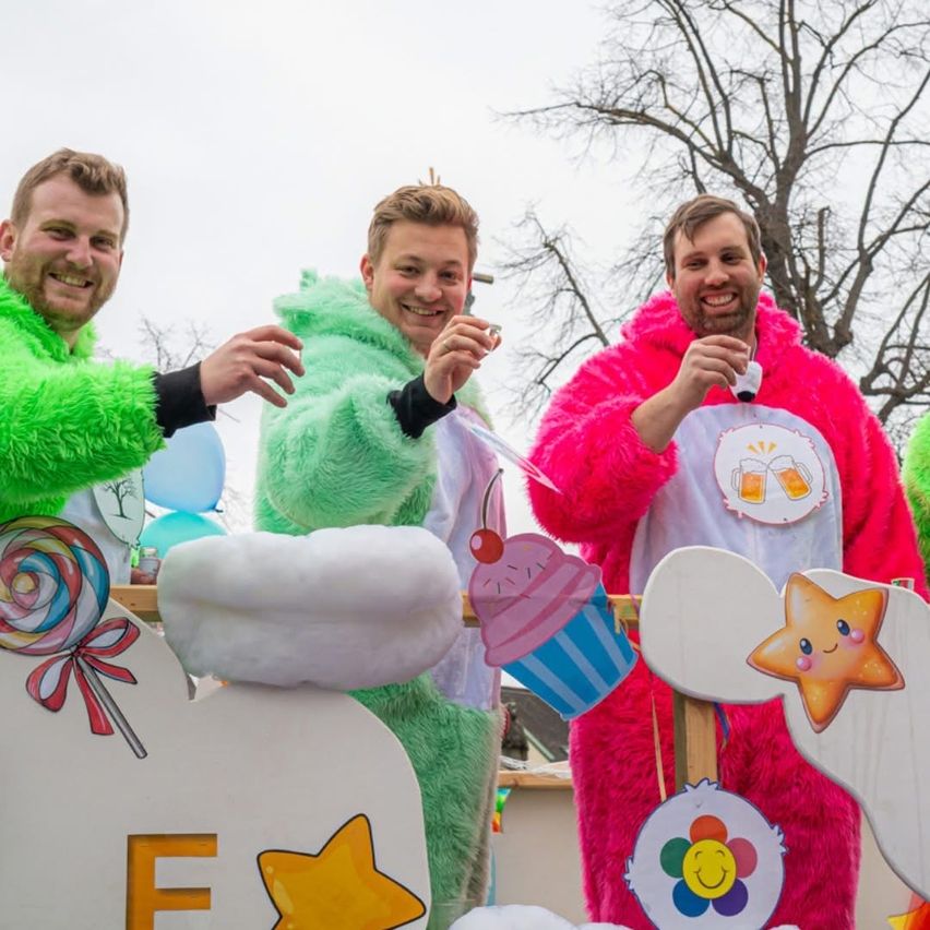 Drei Männer in leuchtenden Kostümen, grün, pink und weiß, stehen lächelnd zusammen. Sie halten Requisiten wie Lollipops und Cupcakes. Ein weißer Hintergrund zeigt bunte Sterne und den Buchstaben F. Dahinter sind Luftballons und ein Baum zu sehen.