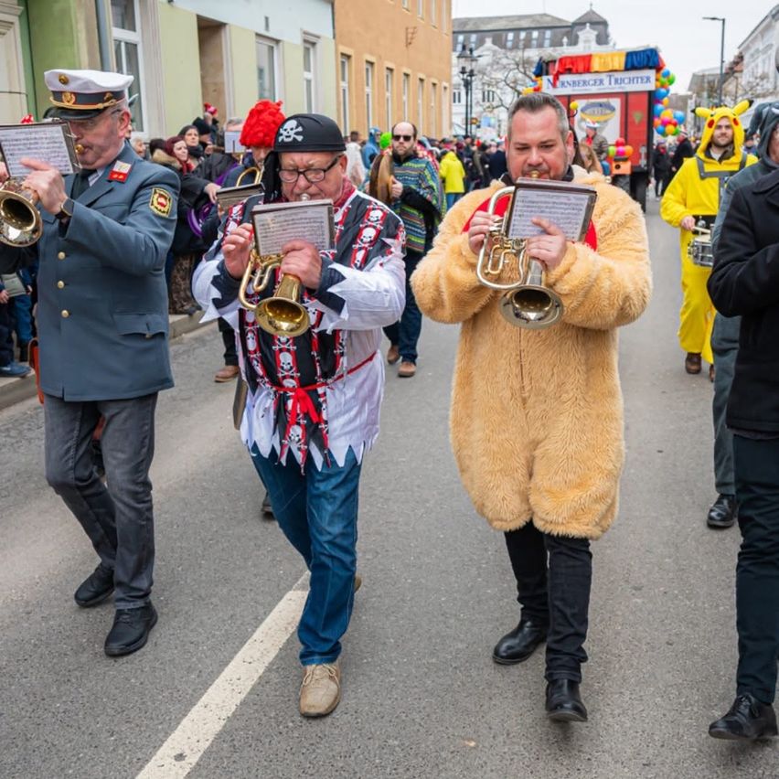 Zwei Männer in Kostümen spielen Trompeten, während sie in einer Straße marschieren. Einer trägt ein Piratenkostüm und der andere ein Bärenkostüm. Hinter ihnen sind mehrere Menschen in Kostümen und ein Umzugswagen.