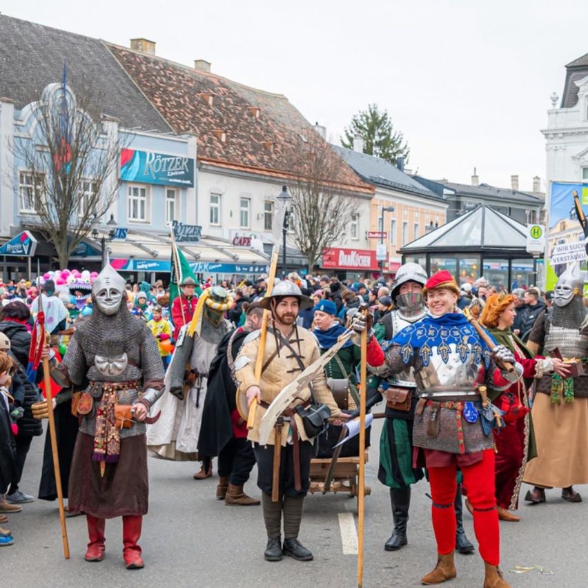 Eine Gruppe von Menschen in mittelalterlichen Kostümen zieht durch eine Straße. Sie tragen Waffen und Banner, mit Gebäuden und Bäumen im Hintergrund.