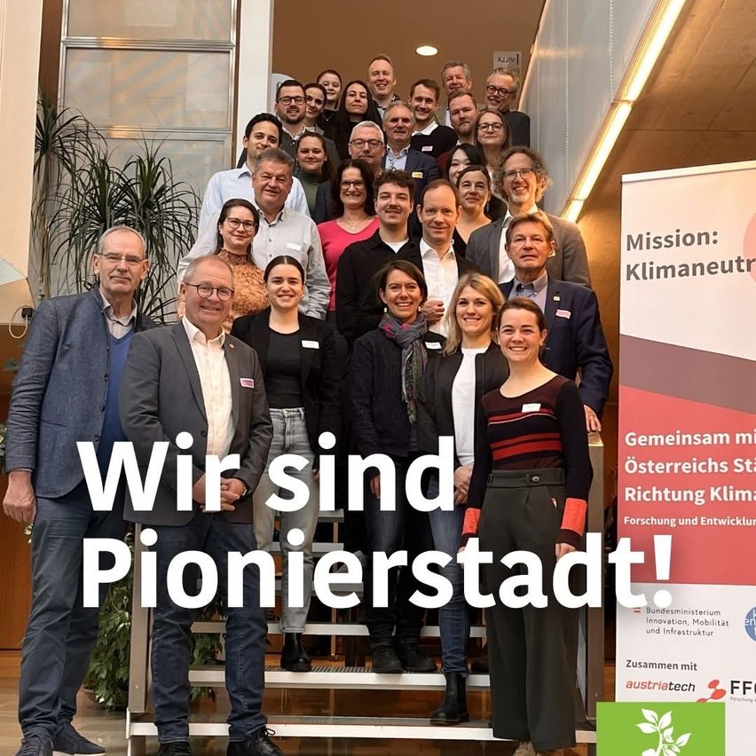Eine Gruppe von Menschen steht auf einer Treppe, lächelt, mit einem Schild 'Wir sind Pionierstadt!' und einem Plakat mit der Überschrift 'Mission: Klimaneutral'.