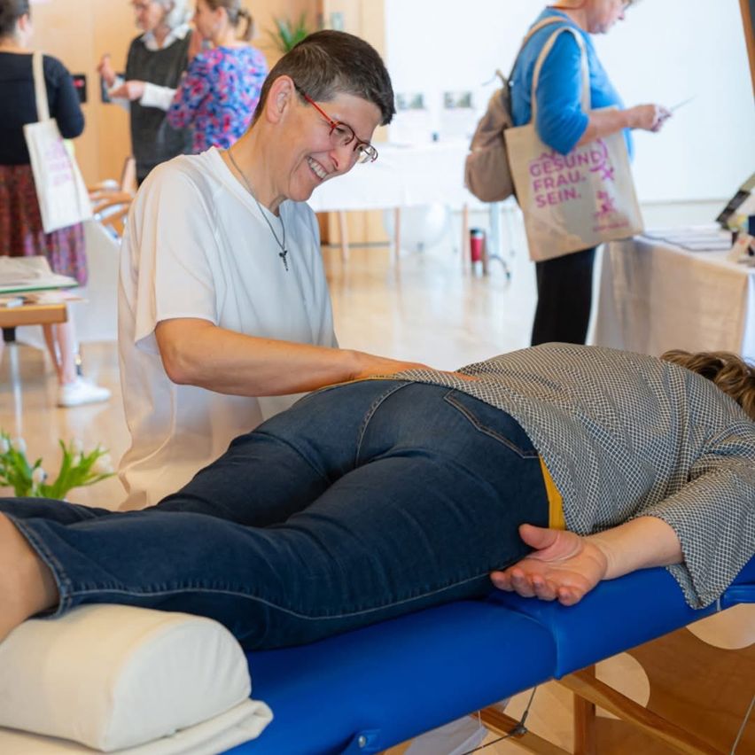 Eine Frau mit Brille liegt auf einem blauen Tisch, während eine andere Frau lächelt und sie berührt. Im Hintergrund stehen zwei Personen.