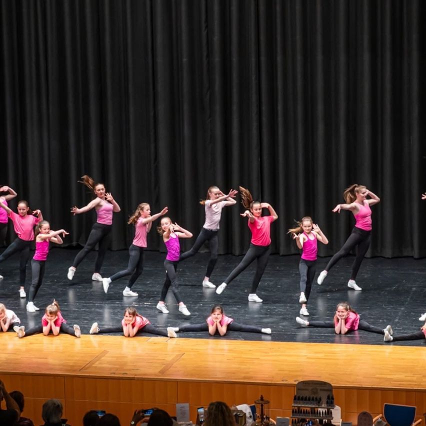 Eine Gruppe junger Mädchen führt eine Tanzroutine auf der Bühne in rosa Shirts und schwarzen Leggings auf. Sie sind in Bewegung eingefangen, einige mit erhobenen Armen. Ein schwarzer Vorhang als Hintergrund und zuschauendes Publikum sind sichtbar.