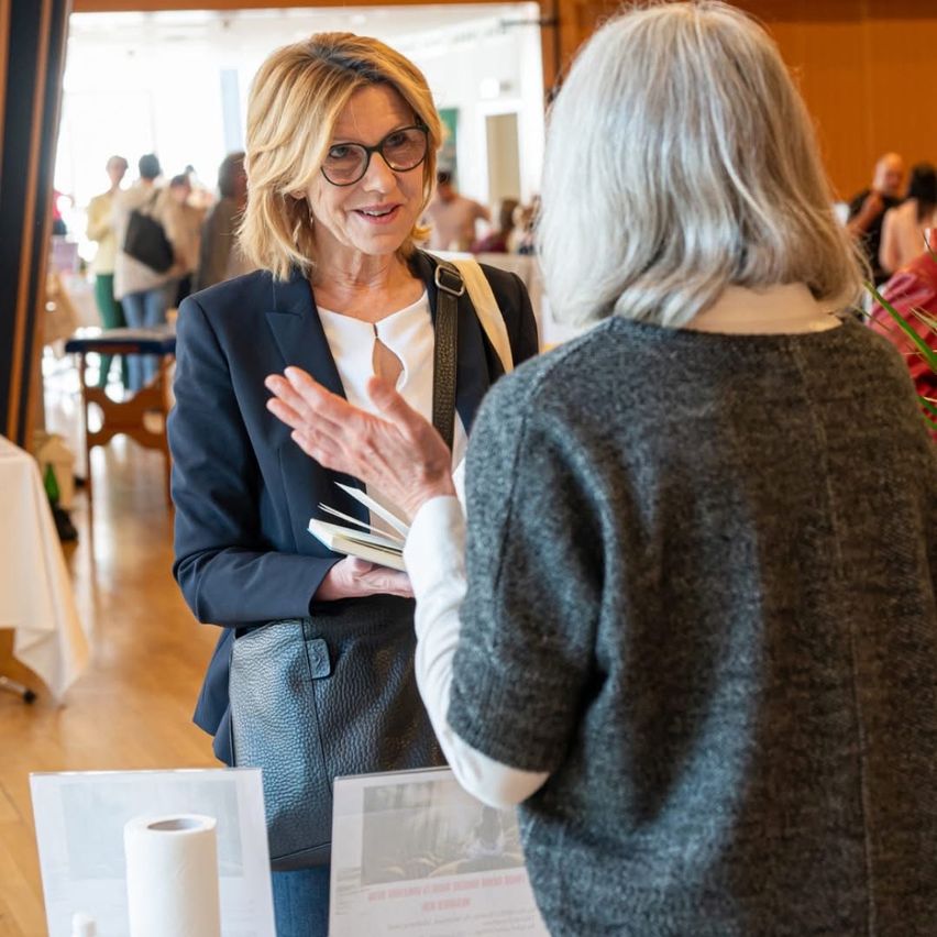Zwei Frauen unterhalten sich bei einer Veranstaltung. Die Frau auf der linken Seite hält ein Buch und trägt eine Brille. Die Frau auf der rechten Seite hört aufmerksam zu. Im Hintergrund ist ein geschäftiger Bereich mit Menschen und Tischen zu sehen.