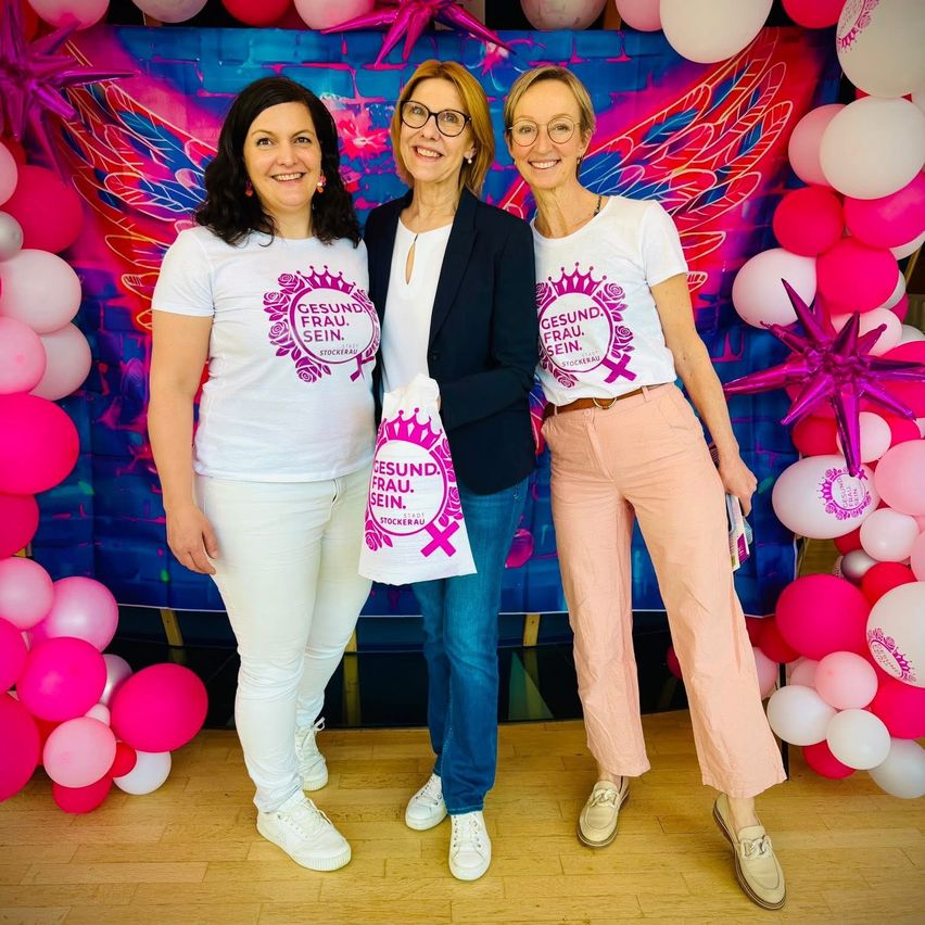 Drei Frauen stehen gemeinsam vor einem farbenfrohen Hintergrund mit Luftballons. Jede trägt ein weißes T-Shirt mit 'Gesund Frau Sein' darauf. Die mittlere Frau hält eine Tasche.