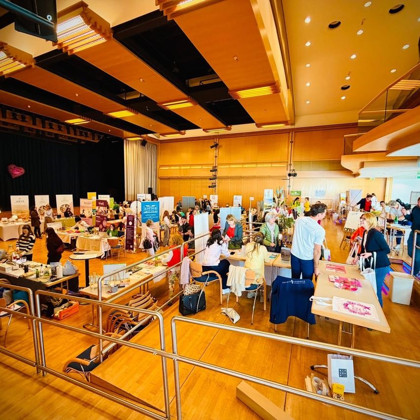 Ein großer Innenraum, der einem Konferenzsaal ähnelt, gefüllt mit Tischen und Stühlen. Menschen sind in verschiedenen Aktivitäten beschäftigt, einige stöbern auf Tischen mit ausgestellten Gegenständen. Die Tische haben Gegenstände und einige haben Schilder. Der Raum hat einen Holzboden, eine Bühne und eine Decke mit Lichtern.