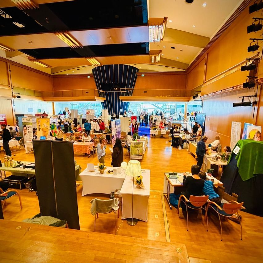 Eine geschäftige Indoor-Veranstaltung mit mehreren Ständen, Tischen und Stühlen. Die Leute unterhalten sich und plaudern. Der Raum hat Holzböden und -wände, mit Lampen und Postern.