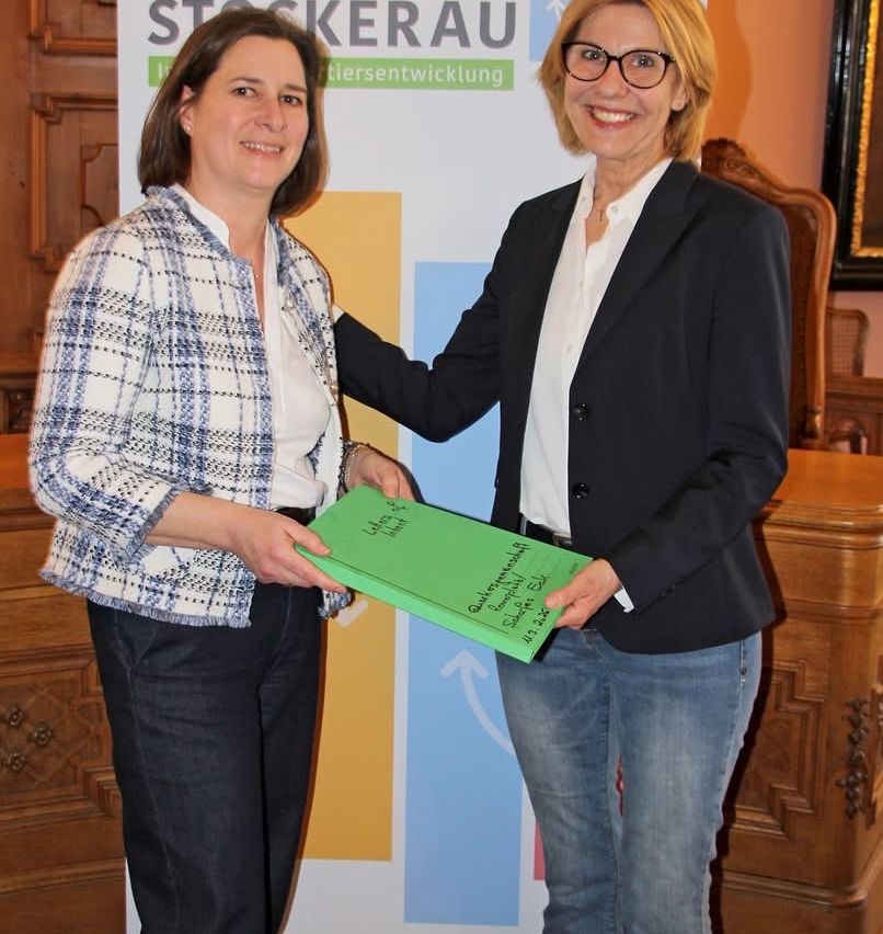 Zwei Frauen stehen nebeneinander und halten ein grünes Büchlein vor einem farbenfrohen Hintergrund. Die Frau links trägt einen karierten Blazer.