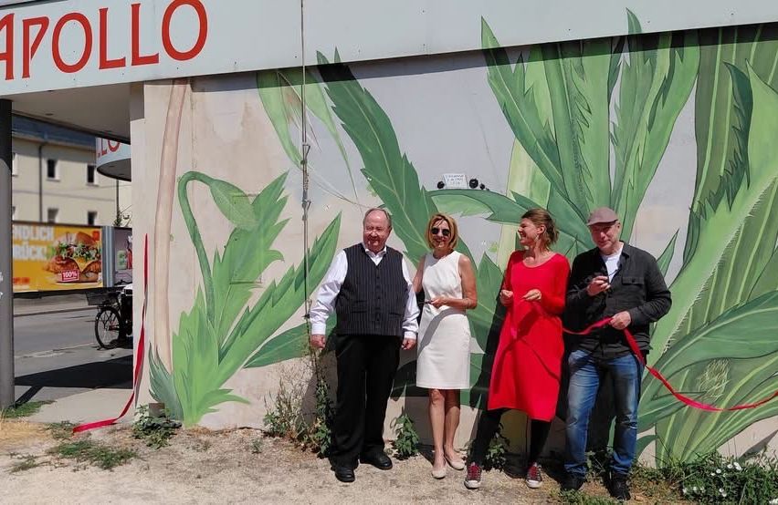 Vier Personen stehen vor einer Wand mit einem Wandbild aus grünen Blättern und einem Band. Der Mann auf der linken Seite trägt eine Weste, und die Frau in der Mitte trägt ein weißes Kleid. Der Mann auf der rechten Seite hält ein Band. Hinter ihnen steht ein Gebäude mit dem Wort 'POLLO' oben.