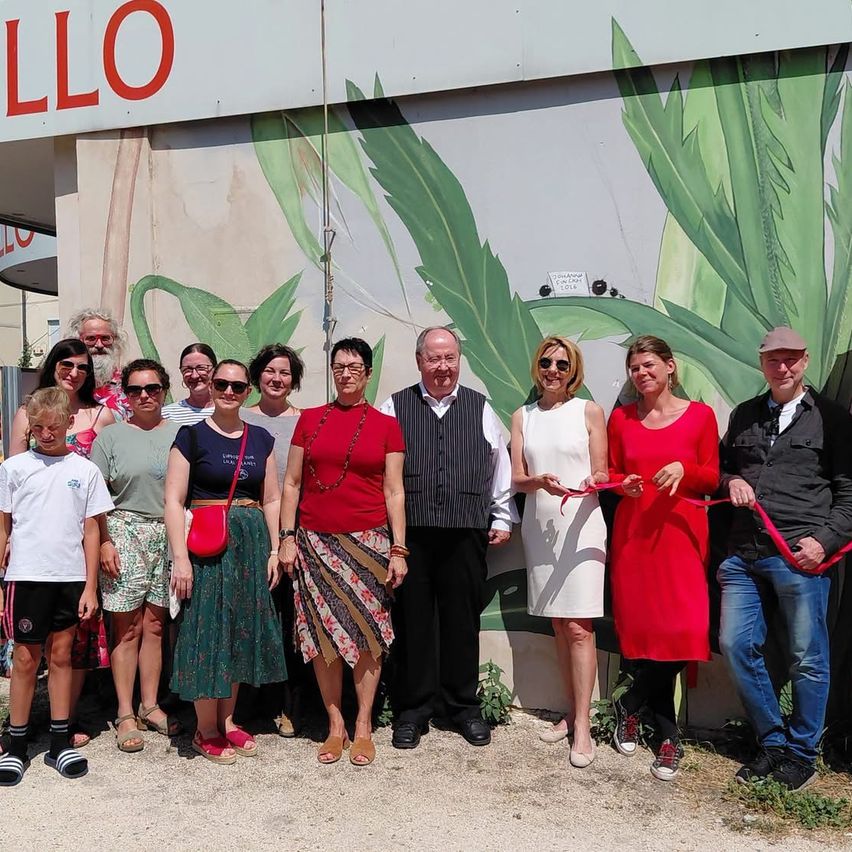 Eine Gruppe von Menschen steht vor einem Gebäude mit einem grünen Blatt-Mural und hält ein rotes Band für eine Bandschneidezeremonie.