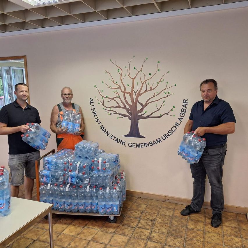 Drei Männer stehen in einem Raum und halten Kisten mit Wasser in der Hand, mit einem großen Baumwandbild hinter ihnen.