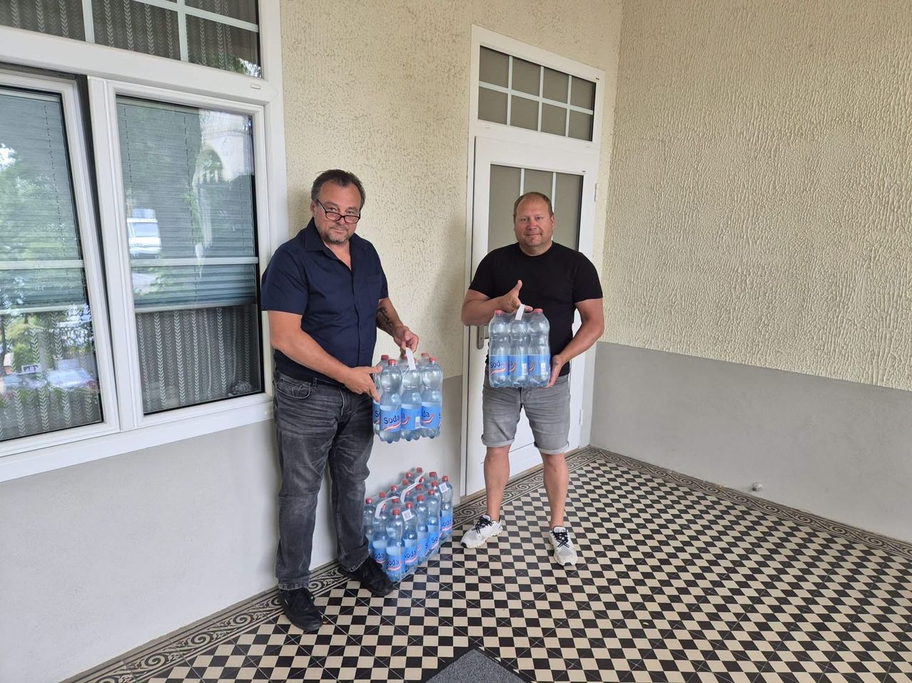 Zwei Männer stehen in einem Raum mit einem karierten Boden und halten Wasserflaschen. Ein Mann trägt ein blaues Hemd und Jeans, der andere ein schwarzes Hemd und Shorts. Sie lächeln beide und scheinen für ein Foto zu posieren.