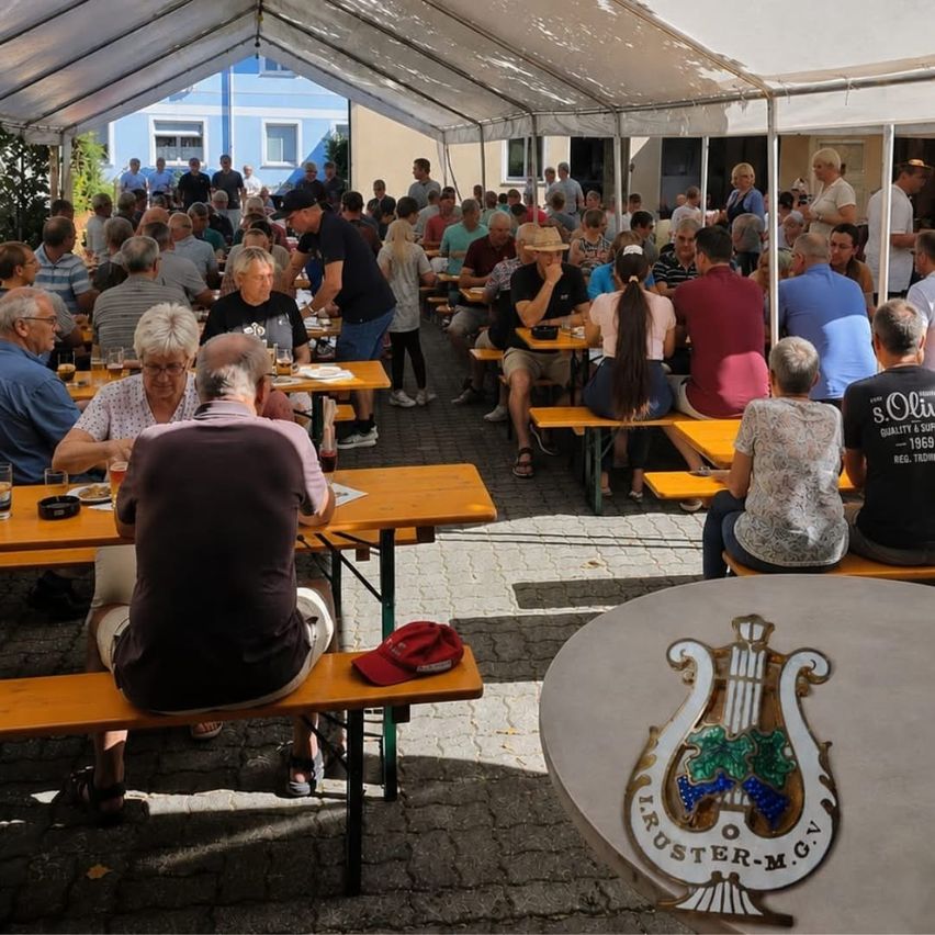 Ein Freiluft-Essbereich unter einem Zelt ist voll mit Menschen. Viele sitzen an Holzbänken um Tische herum, während andere stehen und sich unterhalten. Ein Tisch im Vordergrund zeigt einen roten Hut und eine Brille. Im Hintergrund steht ein Gebäude mit blauen Wänden.