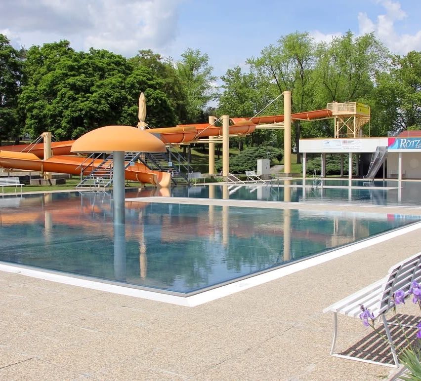 Ein Wasserpark mit großem Pool, einem pilzförmigen Sonnenschirm und orangefarbenen Rutschen. Bäume und ein Gebäude im Hintergrund.