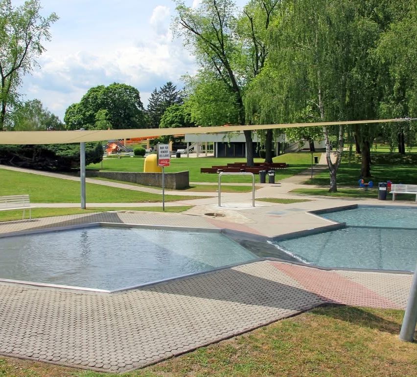 Ein Freibadbereich mit einer Bank und einem Schild. Zwei Becken, eines mit einer Wasserrutsche, und ein Rasenbereich mit Bäumen.