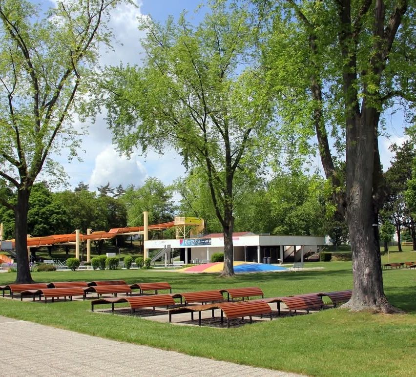 Ein Graspark mit mehreren Holzbänken, einem großen Gebäude mit einer Rutsche im Hintergrund und Bäumen mit üppigen grünen Blättern.