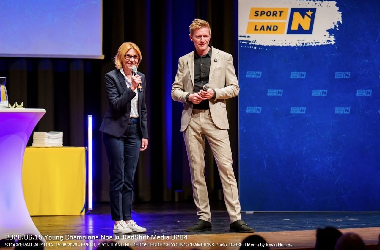 Eine Frau und ein Mann stehen auf einer Bühne und halten Mikrofone. Die Frau trägt eine Brille, einen Blazer und Hosen. Der Mann trägt einen Anzug und Schuhe. Hinter ihnen befindet sich ein Hintergrund mit dem Text 'SPORT LAND N'.