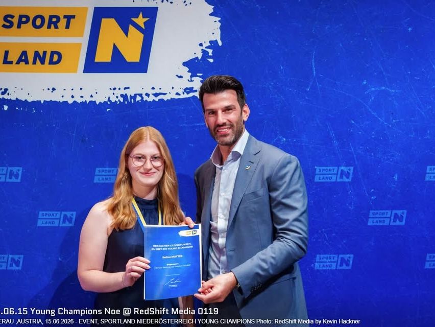 Ein Mann und eine Frau stehen vor einem blauen Hintergrund mit einem Logo. Die Frau hält ein Zertifikat und lächelt. Der Mann, in einem Anzug gekleidet, hält das Zertifikat mit einem Stift. Der Hintergrund hat den Text 'SPORT LAND N'.
