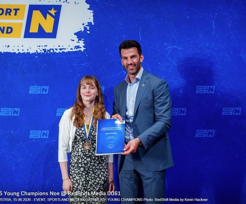 Ein Mann und eine Frau stehen vor einem blauen Hintergrund mit Logos. Sie halten ein blaues Zertifikat. Die Frau trägt eine Halskette und ein Armband. Der Mann trägt einen Anzug und eine Krawatte. Beide lächeln für die Kamera. Foto von RedShift Media.