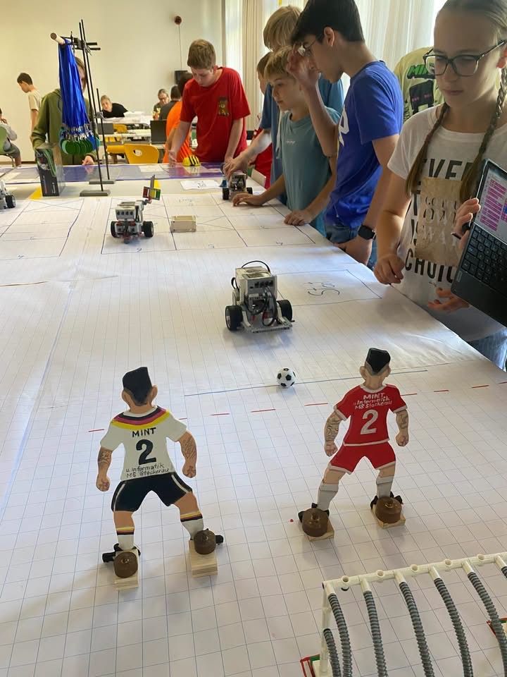 Eine Gruppe von Kindern in verschieden farbigen Shirts beobachten zwei kleine Fußballfiguren auf einem Rastermuster-Boden mit Robotern darauf.