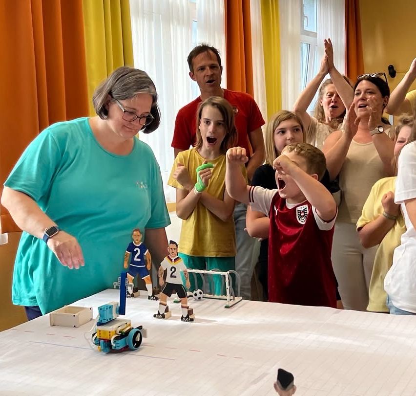 Eine Gruppe von Kindern und Erwachsenen versammelt sich um einen Tisch und schaut sich ein Roboter-Fußballspiel an. Sie jubeln und sind aufgeregt. Das Spiel ist auf einer weißen Fläche mit Spielzeugfiguren.