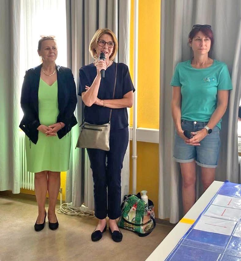 Drei Frauen stehen in einem Raum, eine mit einem Mikrofon, wahrscheinlich bei einer Präsentation. Hinter ihnen ein gelber Wand, ein weißer Vorhang und ein Tisch mit einer blauen Abdeckung.