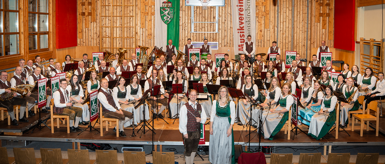 Eine große Gruppe von Musikern, sowohl Männer als auch Frauen, gekleidet in traditioneller Tracht, tritt auf einer Bühne auf, die mit einer grün-weißen Flagge im Hintergrund geschmückt ist.