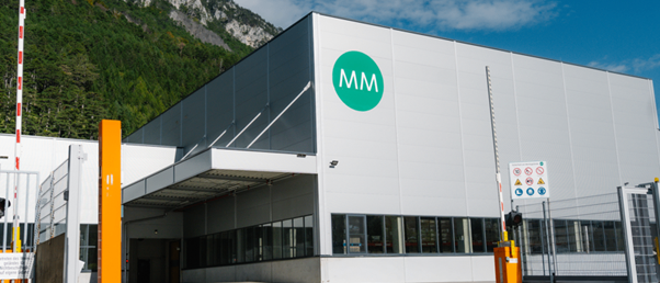 Ein großes weißes Industriegebäude mit einem grünen kreisförmigen Logo mit 'MM' steht vor dem Hintergrund von Bergen. Das Gebäude hat ein modernes Design mit großen Fenstern und einem modernen Tor.